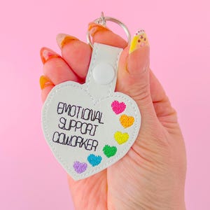 May include: White heart-shaped keychain with the text "Emotional Support Coworker" and rainbow-colored hearts.
