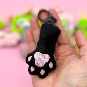 May include: A black cat paw keyring with pink embroidered paw pads. The keyring has a black clip and is held in a hand against a blurred background of other plush toys. The paw is made of soft material.