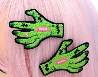May include: Two green zombie hand hair clips with pink accents. The hands are embroidered with black stitching and have a distressed look.