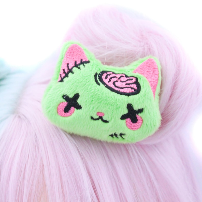 May include: Green plush cat-shaped hair clip with pink ears and a pink brain design. The cat has black eyes with an X over them and a pink nose.