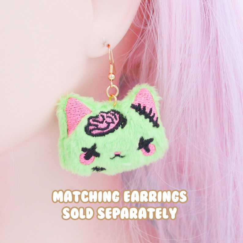 May include: Green fuzzy cat-shaped earrings with pink details and a black embroidered brain. The earrings are hanging from gold hooks. "Matching earrings sold separately" is written below the earrings.