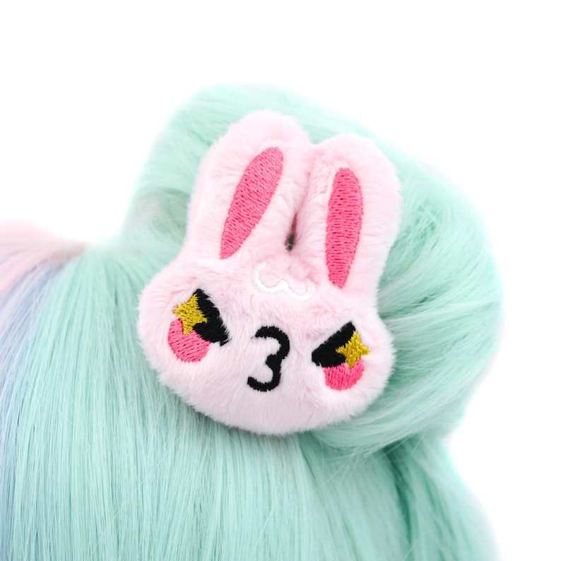 Pu&ograve; includere: Un fermaglio per capelli in peluche rosa a forma di coniglio con occhi ricamati, il numero "3" e stelle gialle. Il coniglio ha un'espressione arrabbiata.