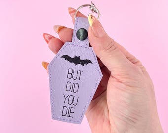 Puede incluir: Un llavero en forma de ataúd de color morado claro con texto bordado en negro que dice BUT DID YOU DIE y un gráfico de murciélago negro. El llavero cuenta con un cierre de botón a presión oscuro y una anilla metálica plateada.