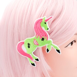 Puede incluir: Una pinza para el pelo de unicornio zombie verde y rosa. El unicornio est&aacute; de pie sobre sus patas traseras con las patas delanteras levantadas. El unicornio tiene una melena y una cola rosas.