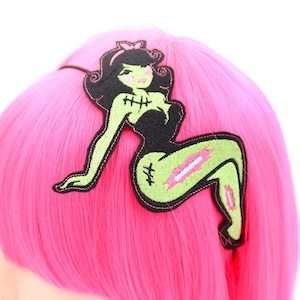May include: A black headband featuring an embroidered patch of a green-skinned figure with black hair, wearing a black outfit. The figure has stitched details on the chest and legs, with pink accents on the cheeks and clothing tears.