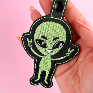 以下が含まれることがあります： 黒い布製のキーホルダーに、黒い目をした緑色のエイリアンが刺繍されています。エイリアンの両手にはピースサインが刺繍されています。