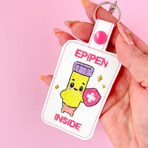 Könnte beinhalten: Weißer Schlüsselanhänger mit rosa Nähten und einem Cartoon-Epipen mit einem rosa Kreuz auf seinem Schild. Der Text "EPIPEN INSIDE" ist auf den Schlüsselanhänger gestickt.