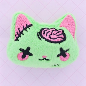 May include: Green plush cat with pink ears and eyes, stitched closed with black X's. The cat has a pink brain embroidered on its head.