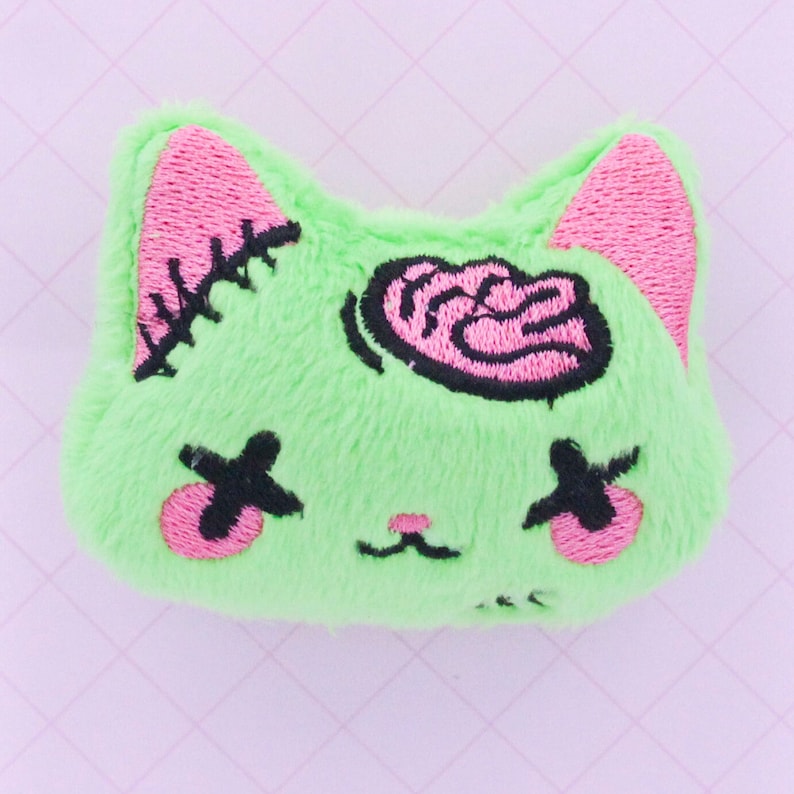 May include: Green plush cat with pink ears and eyes, stitched closed with black X's. The cat has a pink brain embroidered on its head.