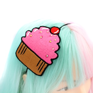 May include: A pink cupcake headband with a red cherry on top. The cupcake is decorated with white icing and sprinkles. The headband is black.