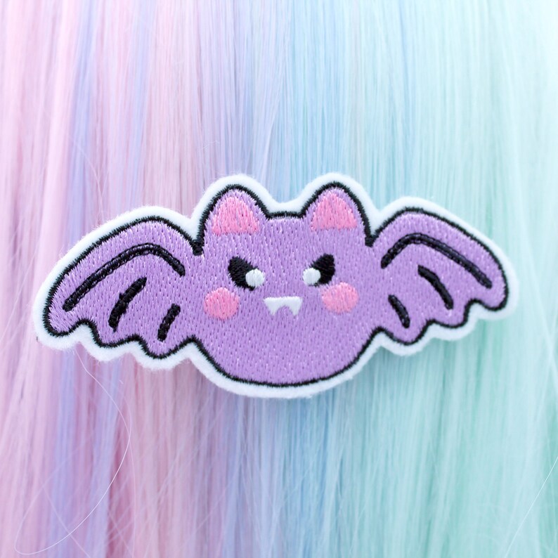May include: Purple embroidered iron-on patch of a cartoon bat with a grumpy expression. The bat has black eyes, white teeth, and pink cheeks.