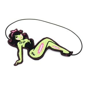 May include: An embroidered patch featuring a stylized green-skinned figure with black hair, a pink bow, and black clothing. The figure has visible stitches on the chest and leg, with pink accents on the skin, attached to a thin black elastic cord.