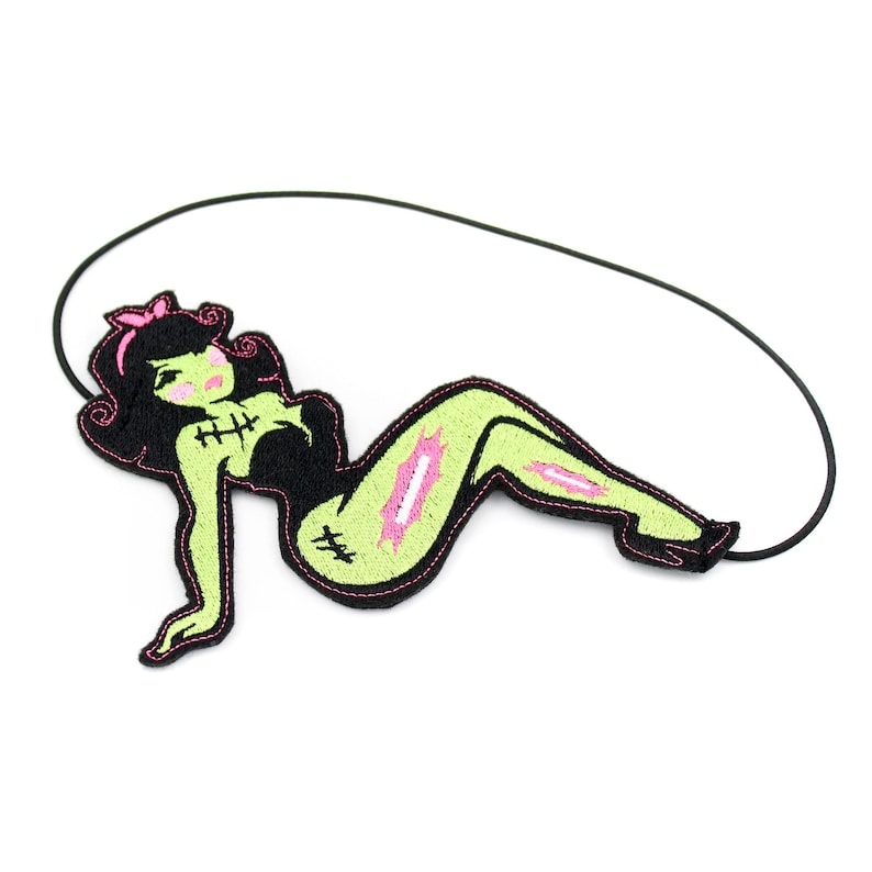 May include: An embroidered patch featuring a stylized green-skinned figure with black hair, a pink bow, and black clothing. The figure has visible stitches on the chest and leg, with pink accents on the skin, attached to a thin black elastic cord.
