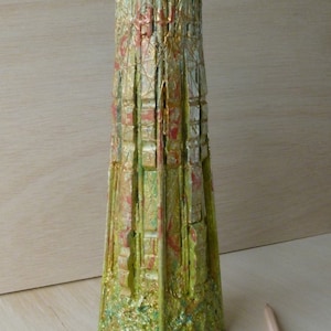 Può includere: Un vaso decorativo e strutturato con una forma architettonica distintiva. Il vaso presenta tonalità verdi, dorate e rosse, con una texture ruvida e stratificata. Il vaso è alto circa 30 cm. Due matite sono in primo piano.