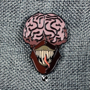 May include: A pink and brown enamel pin depicting a brain with a toothy, open mouth and a pink tongue. The brain is stylized to look like a monster.