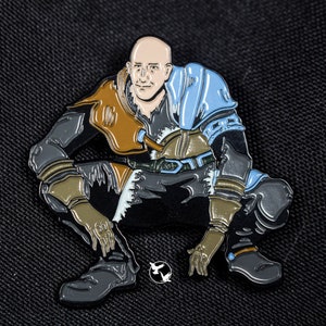 May include: A black and brown enamel pin depicting a bald man in a crouching pose, wearing a brown and blue outfit with a blue cape. The pin has a detailed design and a glossy finish.