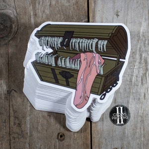 May include: A stack of stickers featuring a cartoon illustration of a treasure chest with a tongue sticking out. The chest is brown with gold accents and the tongue is pink.