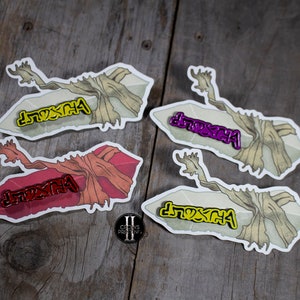 May include: Four enamel pins with a stylized design of a tree branch and the word "Dream" in different colors: yellow, purple, red, and yellow. The pins are on a wooden surface.