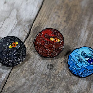 Könnte beinhalten: Drei runde Pins mit einer strukturierten Oberfläche und einem einzelnen Auge in der Mitte. Die Augen sind in verschiedenen Farben: rot, schwarz und blau.