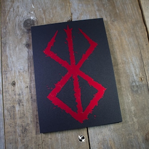 May include: A black notebook with a red, stylized symbol on the cover. The symbol has a geometric design with sharp angles and a textured, splatter-like effect. The notebook is on a wooden surface.