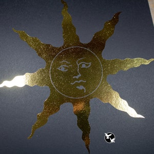 May include: A gold foil print of a sun with a face on a black background.