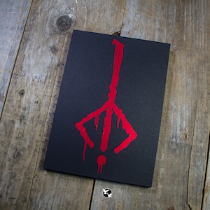 May include: A black rectangular card featuring a red, stylized symbol. The symbol has a vertical line with a three-pronged base and a small circle at the bottom. The card is on a wooden surface.