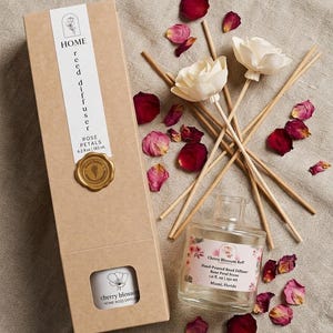 May include: A reed diffuser set featuring a tall tan cardboard box labeled HOME reed diffuser Rose Petals 6.2 fl oz, a clear glass bottle with a Cherry Blossom Reed label, and two white wooden flower-topped reeds surrounded by red rose petals.