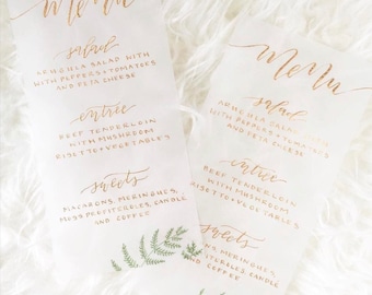 Może przedstawiać: Dwa złote menu kaligraficzne na białym papierze z zielonymi ilustracjami paproci. Menu zawiera sałatkę, danie główne i desery na wesele lub imprezę. Tekst na menu brzmi: "Sałatka: Sałatka z rukoli z papryką + pomidorami i serem feta", "Danie główne: Polędwica wołowa z grzybami Risotto + warzywa", i "Desery: Makaroniki, bezy, profiterolki z kremem, świeca i kawa".