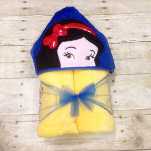 May include: A yellow hooded towel with a blue hood featuring an embroidered image of Snow White. The towel has a blue ribbon tied around it.