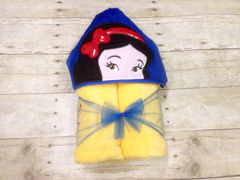 May include: A yellow hooded towel with a blue hood featuring an embroidered image of Snow White. The towel has a blue ribbon tied around it.
