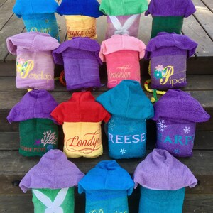 May include: A collection of 14 hooded towels in various colors, each with a personalized name embroidered on the back. The towels are arranged in a staircase pattern on a wooden surface.