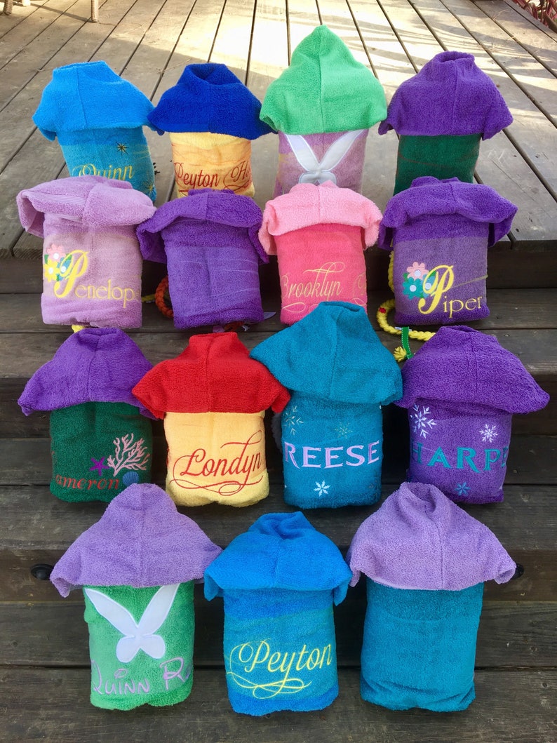 May include: A collection of 14 hooded towels in various colors, each with a personalized name embroidered on the back. The towels are arranged in a staircase pattern on a wooden surface.