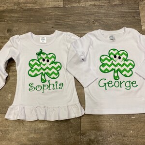 May include: Two white long-sleeved shirts with green shamrock designs and personalized names. The shirt on the left says "Sophia" and the shirt on the right says "George".