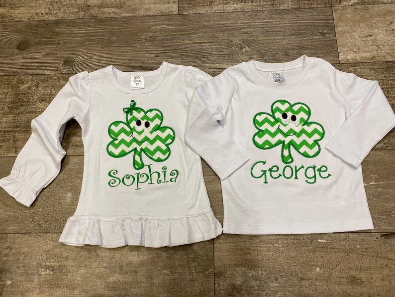 May include: Two white long-sleeved shirts with green shamrock designs and personalized names. The shirt on the left says "Sophia" and the shirt on the right says "George".