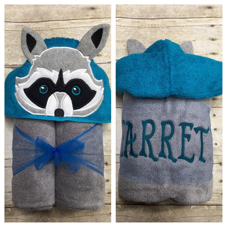 May include: A gray hooded towel with a raccoon face on the hood. The towel is tied with a blue ribbon and the name "Jarret" is embroidered on the back.