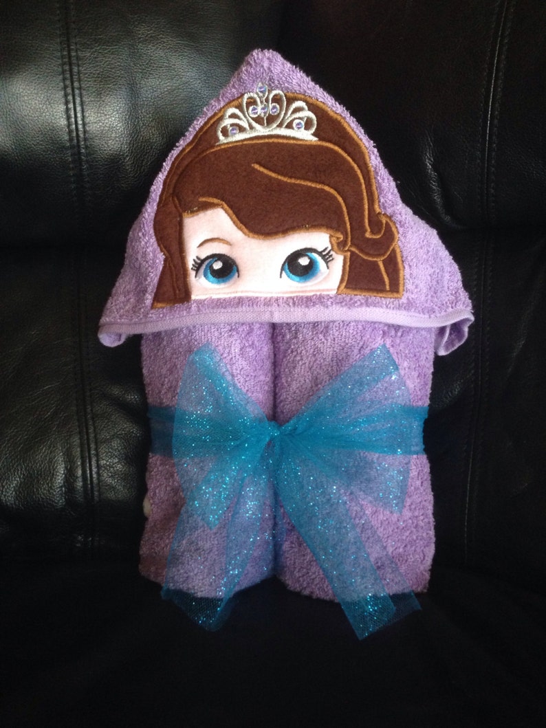 May include: Purple hooded towel with a princess character embroidered on the hood. The towel is tied with a blue sparkly ribbon.