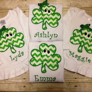 May include: Three white long-sleeved shirts with green shamrock appliques featuring a smiling face and a chevron pattern. Each shirt has a different name embroidered on the bottom: Lyda, Ashlyn, and Maggie. A fourth shirt with the name Emma is shown in the background.