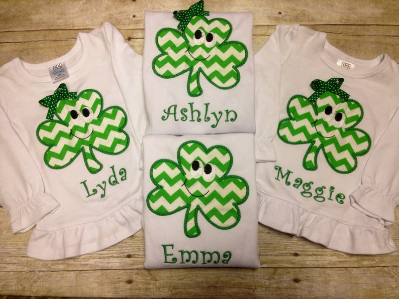 May include: Three white long-sleeved shirts with green shamrock appliques featuring a smiling face and a chevron pattern. Each shirt has a different name embroidered on the bottom: Lyda, Ashlyn, and Maggie. A fourth shirt with the name Emma is shown in the background.