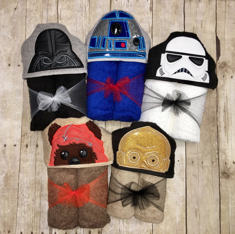 May include: Six hooded towels featuring Star Wars characters. The towels are folded and tied with ribbon. The characters include Darth Vader, R2-D2, Stormtrooper, Chewbacca, and C-3PO.