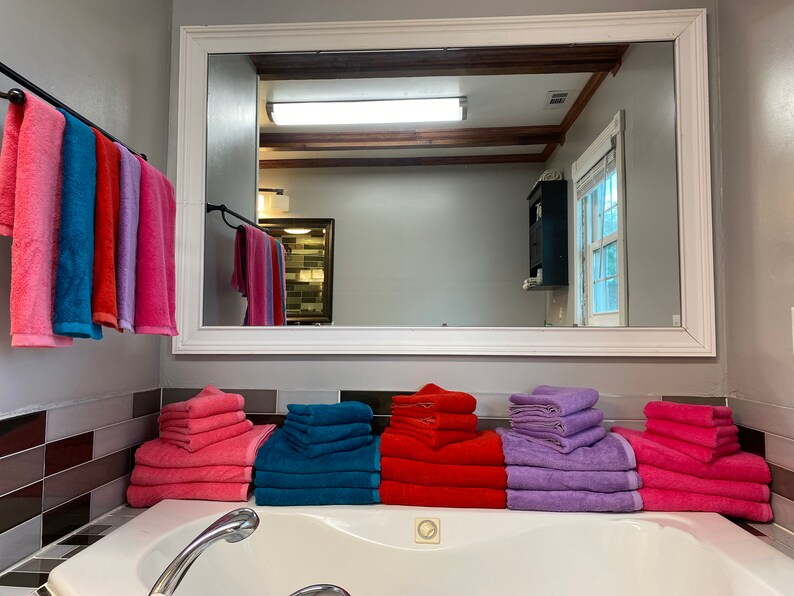 Pu&ograve; includere: Un bagno con una vasca da bagno bianca, uno specchio e un portasciugamani con diversi asciugamani di diversi colori. Ci sono anche pile di asciugamani rosa, viola, rosso e blu sul bancone.