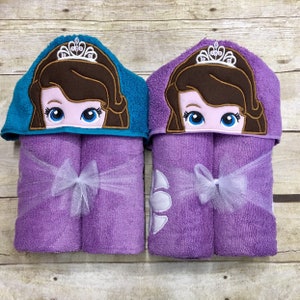 May include: Two purple hooded towels with embroidered princess faces and crowns. The towels are tied with white ribbon bows.