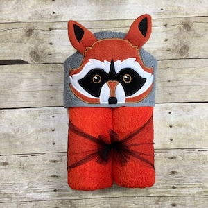Peut inclure: Une serviette &agrave; capuche avec un visage de raton laveur brod&eacute; sur la capuche. La serviette est orange et grise et est attach&eacute;e avec un ruban noir.