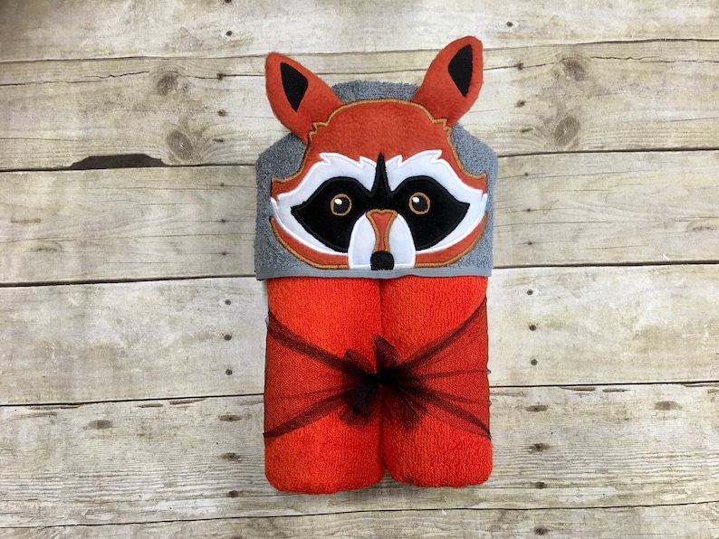 Peut inclure: Une serviette &agrave; capuche avec un visage de raton laveur brod&eacute; sur la capuche. La serviette est orange et grise et est attach&eacute;e avec un ruban noir.