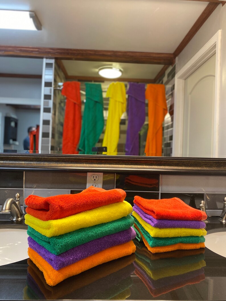 Peut inclure: Une pile de serviettes de bain de couleur arc-en-ciel pli&eacute;es soigneusement sur un comptoir de salle de bain. Les serviettes sont orange, jaune, verte, violette et orange.