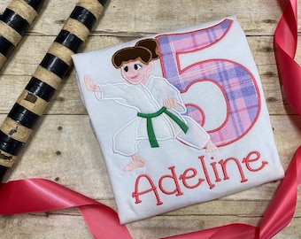 May include: White t-shirt with an embroidered image of a girl in a karate uniform, a pink and purple plaid number "5", and the name "Adeline".
