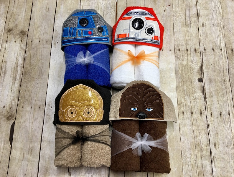 May include: Four hooded towels featuring Star Wars characters. The towels are folded and tied with ribbon. The characters are R2-D2, C-3PO, Chewbacca, and a Stormtrooper.