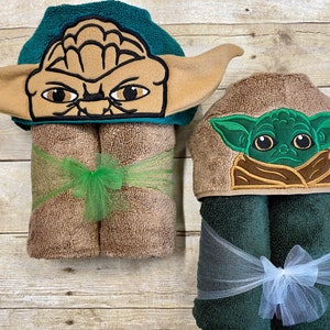 May include: Two hooded towels with embroidered images of Yoda from Star Wars. The larger towel is green and brown with a brown bow. The smaller towel is green and brown with a white bow.