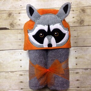 May include: A gray hooded towel with a raccoon face embroidered on the hood. The hood is orange and the towel is gray with an orange bow.
