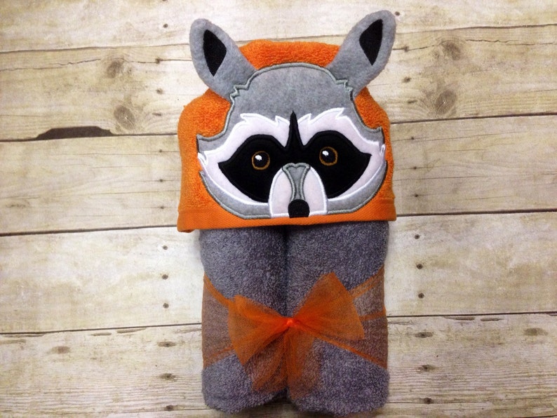 May include: A gray hooded towel with a raccoon face embroidered on the hood. The hood is orange and the towel is gray with an orange bow.