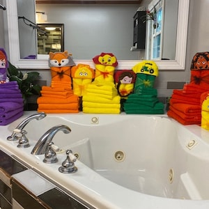 Peut inclure: Une collection de serviettes &agrave; capuche color&eacute;es avec diff&eacute;rents personnages de dessins anim&eacute;s, dont un renard, un lion, une princesse, un tracteur et un oiseau. Les serviettes sont empil&eacute;es dans une salle de bain avec une baignoire blanche.